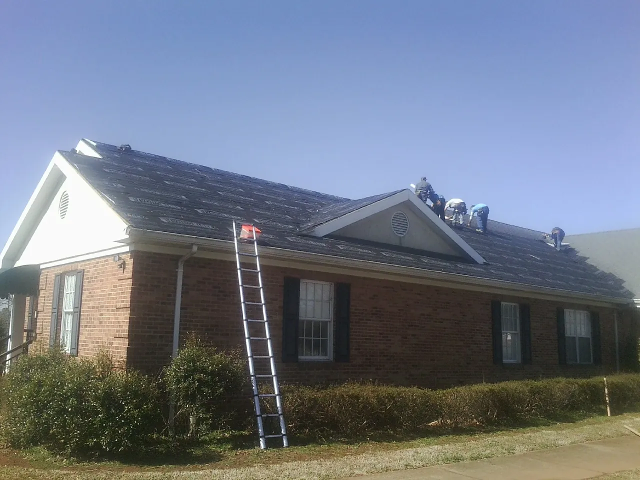 Crew tearing shingles off a brick ranch roof with a ladder set — Metal Roof Repair work by our crew in Andale